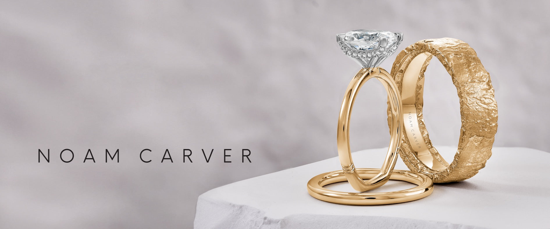 Two gold rings with a diamond on a gray background, featuring the brand 'Noam Carver', a Canadian jewellery designer from Montreal.